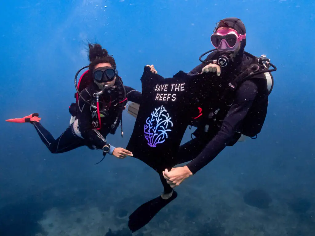 Two divers underwater holding up a black GaiaOne tee printed with Save The Reefs and the coral emblem